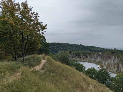 A winding path through grassy hills leads to a river surrounded by trees under a cloudy sky.