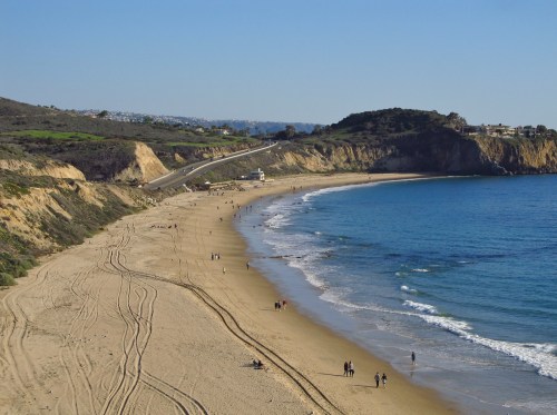 A scenic beach with gentle waves, sandy shore, and people walking along the water, framed by cliffs and greenery.