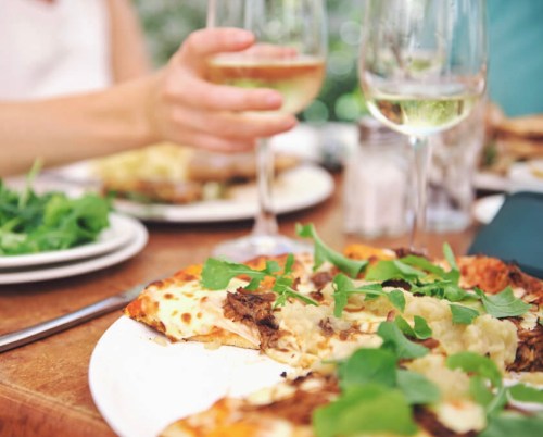 A close-up of a pizza topped with greens, with glasses of white wine and a salad in the background on a wooden table.