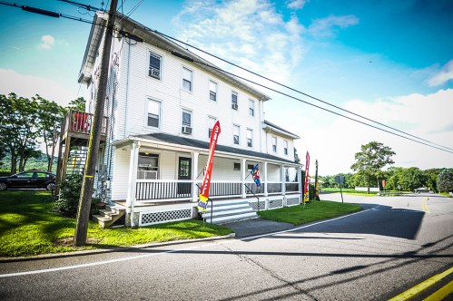 A white multi-story building on a roadside, featuring colorful flags and a grassy area in front.