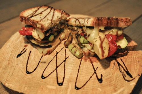 A sliced sandwich with grilled vegetables and melted cheese, drizzled with balsamic glaze, on a wooden surface.