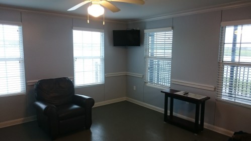 A small room with a leather chair, a table, a TV on the wall, and windows with blinds letting in natural light.