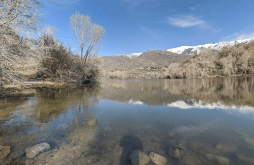 A serene lake surrounded by trees and mountains, with snow-capped peaks under a clear blue sky.