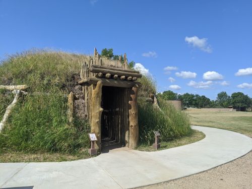 A grassy, earthen structure with a wooden entrance, surrounded by a paved path and open landscape under a blue sky.