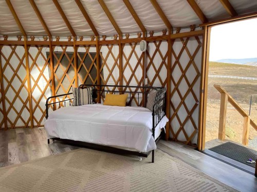 Cozy interior of a yurt featuring a daybed, wooden beams, and a view of the outdoors through an open door.