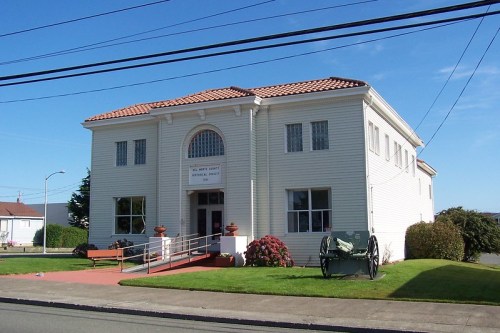 A white building with a red-tiled roof, surrounded by greenery and featuring a historical cannon in front.