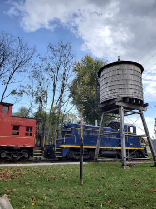 A blue and yellow vintage train next to a red caboose and a wooden water tower, surrounded by trees and a cloudy sky.