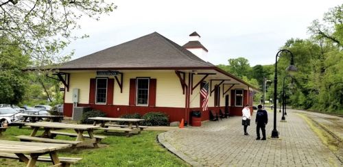 A quaint train station with a red and white exterior, surrounded by picnic tables and greenery, with two people nearby.