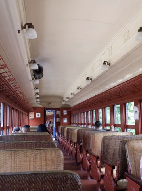 Interior of a vintage train car with rows of upholstered seats and large windows, showing a few passengers.
