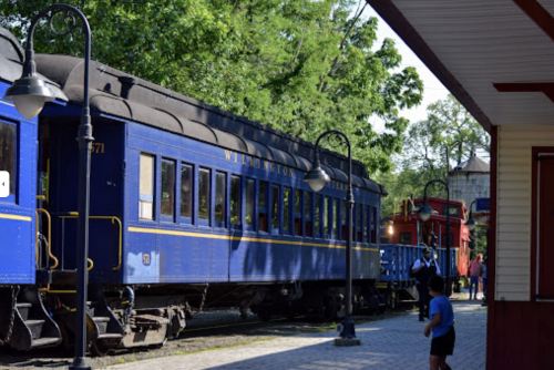 A blue train car at a station, with trees in the background and people nearby.