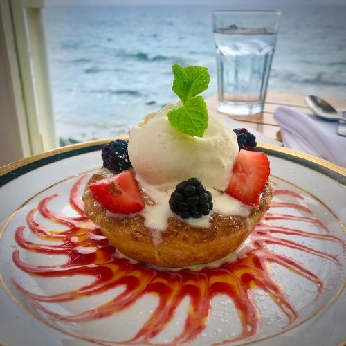 A dessert featuring a pastry bowl filled with ice cream, strawberries, blackberries, and a drizzle of sauce, with an ocean view.