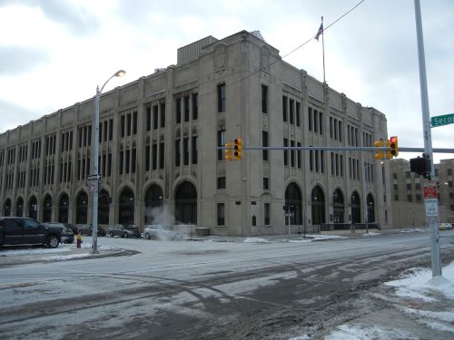 Historic building at a snowy intersection, featuring arched windows and a flag on top, with cars parked nearby.