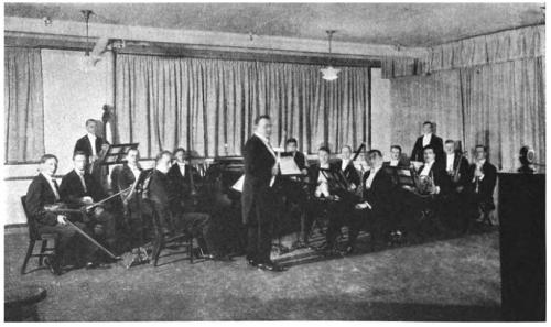 A vintage orchestra poses in a room, with musicians seated and a conductor standing in front of them.