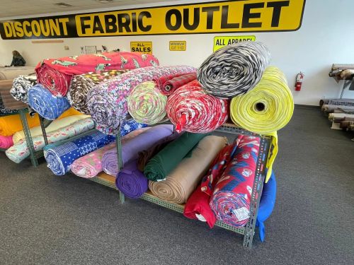 Colorful rolls of fabric stacked on shelves in a discount fabric outlet store, with a large sign overhead.
