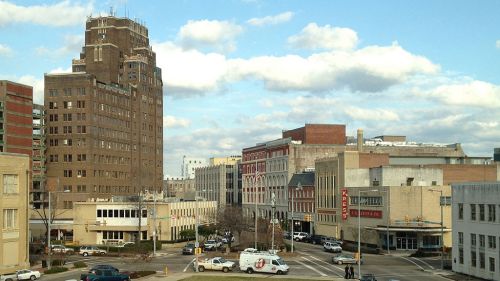 A cityscape featuring tall buildings, a cloudy sky, and busy streets with cars and pedestrians.