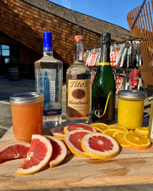 A wooden board displays various drinks: vodka bottles, sparkling wine, and fresh citrus slices in jars.