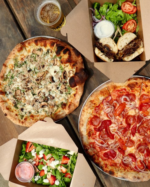A spread of food including two pizzas, a salad with strawberries, and two sandwiches, with a beer in the background.
