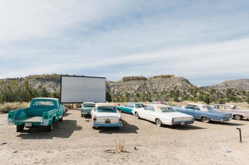 A vintage drive-in theater with classic cars parked in front of a large blank screen and scenic hills in the background.