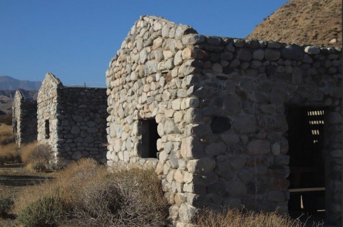 Abandoned stone structures in a dry landscape, surrounded by sparse vegetation and mountains in the background.