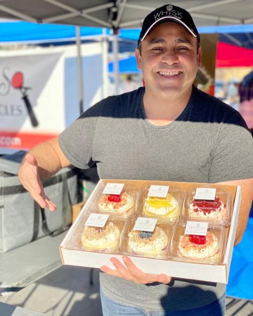 A smiling man holds a box of colorful desserts at a market, with tents in the background.