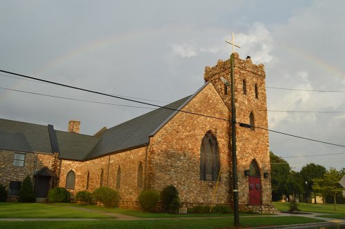 A stone church with a tall tower and cross, under a rainbow and cloudy sky, surrounded by green grass and trees.