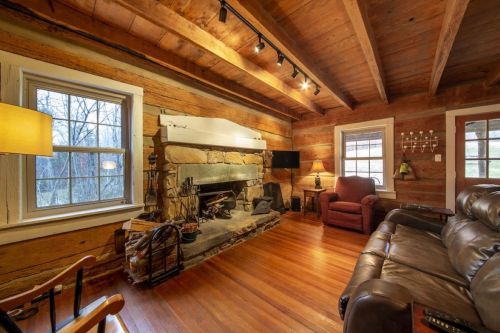 Cozy living room with wooden beams, stone fireplace, and comfortable seating, featuring large windows and warm lighting.