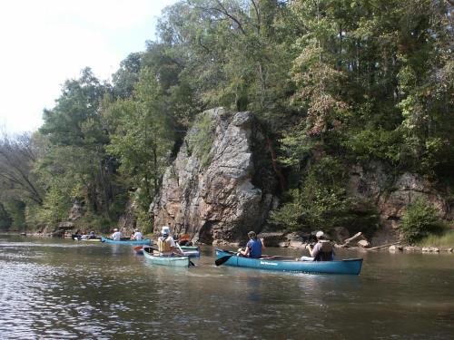 A group of people canoeing on a calm river, surrounded by lush greenery and rocky cliffs.