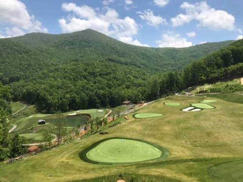 A scenic view of a golf course surrounded by lush green mountains and blue skies with fluffy clouds.