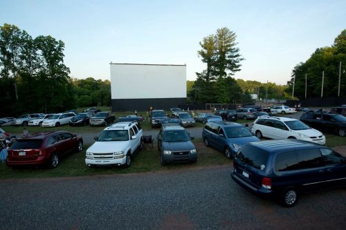 A drive-in movie theater with cars parked in front of a large screen, surrounded by trees and a clear sky.