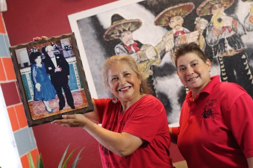 Two women in red shirts smile while holding a framed photo of a couple in formal attire against a colorful background.