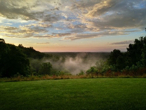 A serene landscape at sunrise, featuring mist rising over a lush green valley and a colorful sky.
