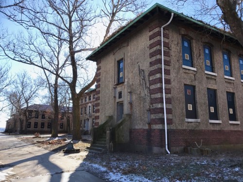 Abandoned building with brick exterior, boarded windows, and bare trees in a desolate landscape. Snow on the ground.