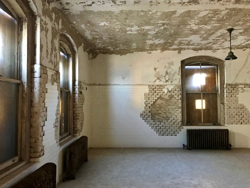 Abandoned room with peeling paint, two windows, and a radiator, featuring exposed brick and tiled walls.
