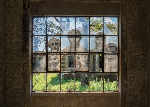 A weathered window with faded portraits of people, framed by greenery outside an abandoned building.