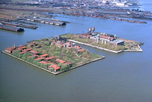 Aerial view of a small island with buildings and greenery, surrounded by water and boats in the background.