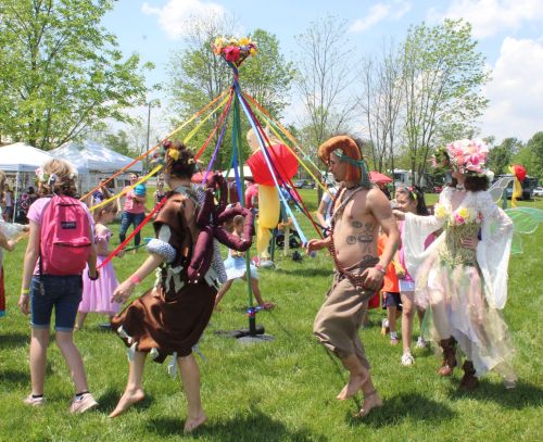 A group of people in costumes dance around a maypole at a festive outdoor event, surrounded by trees and tents.