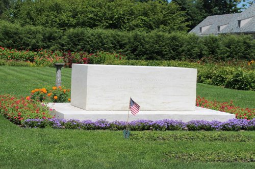 A white stone monument surrounded by colorful flowers and a small American flag in a well-maintained garden.