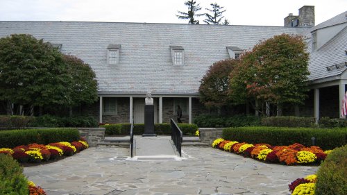 A stone pathway leads to a building with a statue and colorful flower beds in front, surrounded by trees.