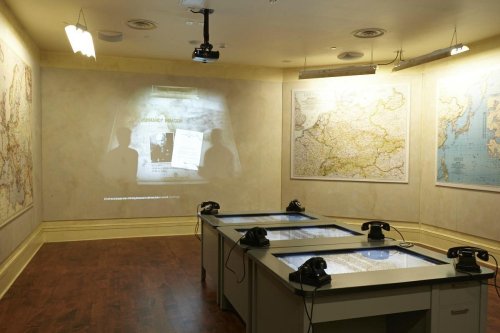 A room with maps on the walls, tables with screens, and vintage telephones, featuring a projected image on the wall.