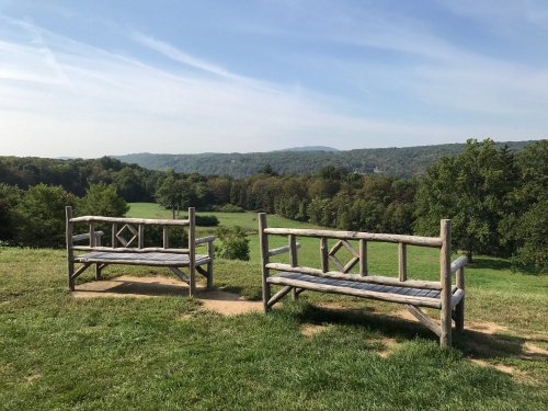 Two wooden benches overlook a lush green landscape with rolling hills under a clear blue sky.