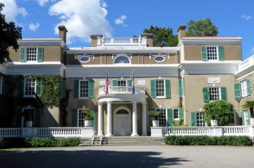 A grand mansion with green shutters, white columns, and flags, surrounded by lush greenery and a clear blue sky.