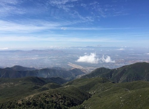 A panoramic view of rolling green mountains under a blue sky with scattered clouds and a distant valley below.