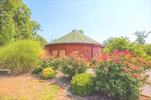 A round log cabin surrounded by colorful flowers and greenery under a clear blue sky.