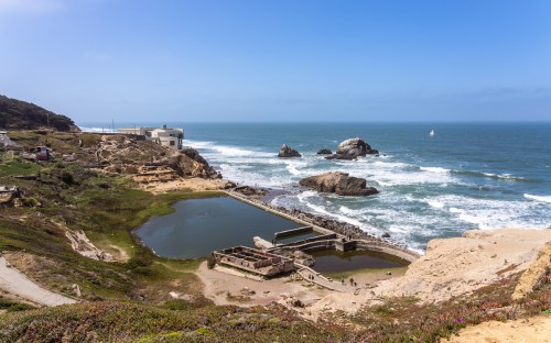 Coastal view featuring rocky outcrops, a calm pond, and waves crashing against the shore under a clear blue sky.