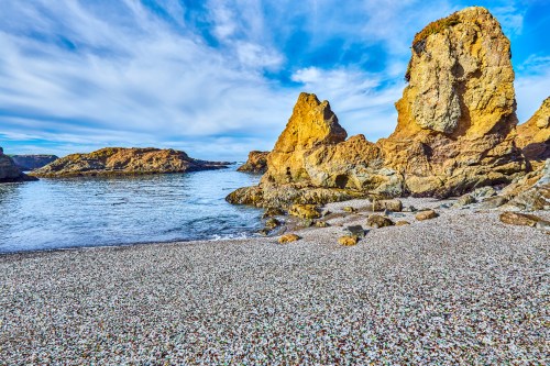 A serene beach scene with rocky formations and calm waters under a blue sky, scattered pebbles on the shore.