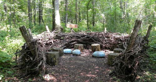 A circular seating area made of branches and logs, surrounded by greenery, with large blue stones in the center.
