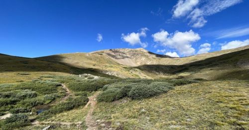A scenic mountain landscape with rolling hills, green vegetation, and a blue sky dotted with clouds.