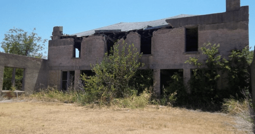 Abandoned building with crumbling walls, broken windows, and overgrown vegetation in a dry landscape.