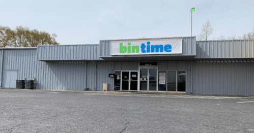 Exterior of a store named "bin time," featuring a large sign and a gray metal facade. Empty parking lot in front.
