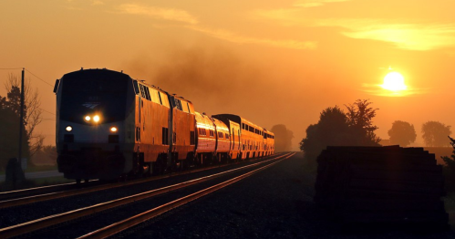 A train travels along tracks at sunset, silhouetted against a vibrant orange sky with trees in the background.